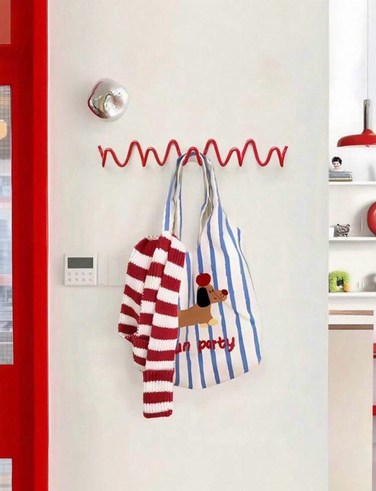 Red wavy wall hook holding a striped tote bag and sweater on white wall.