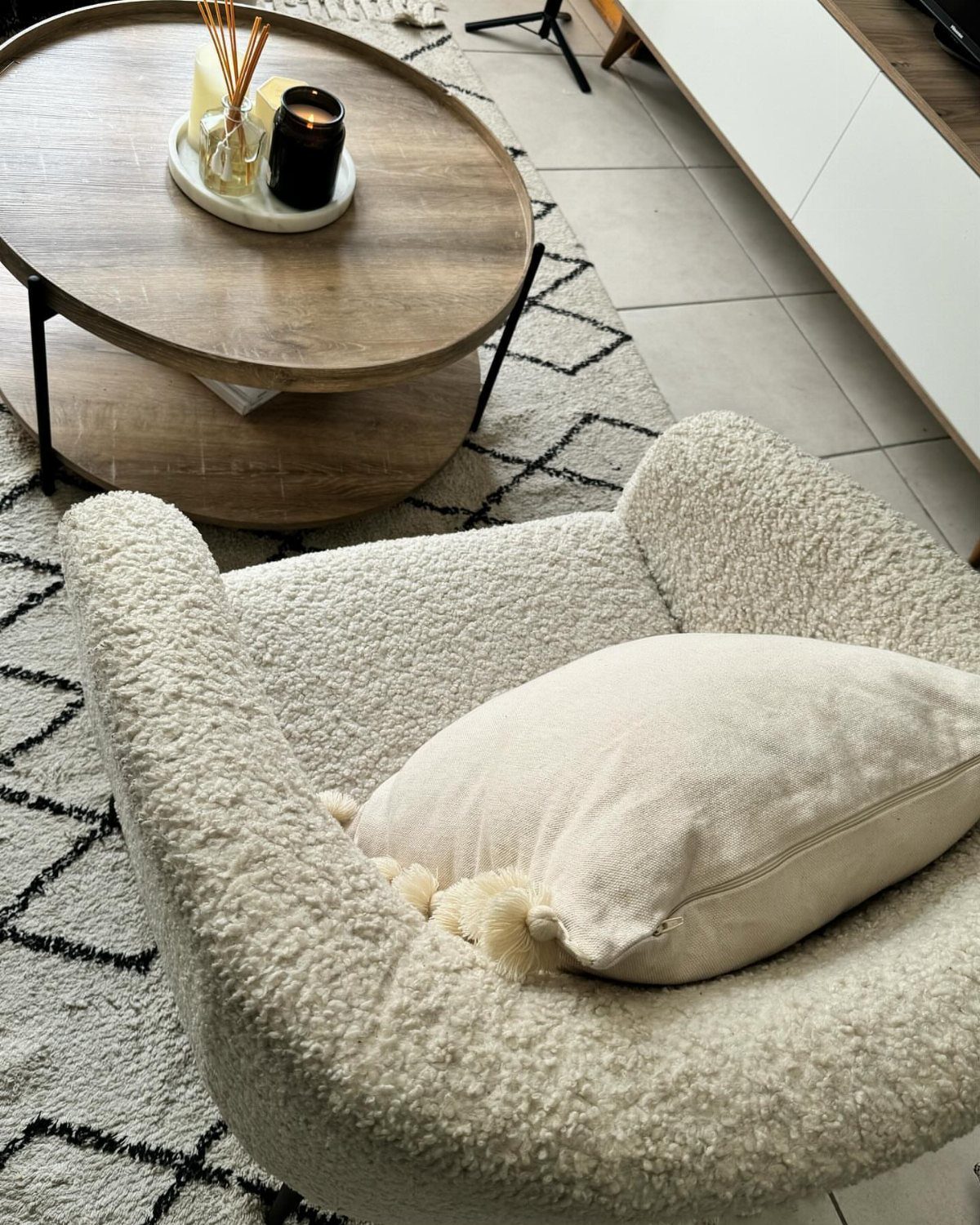 Round wooden coffee table with black metal legs next to a white bouclé armchair.