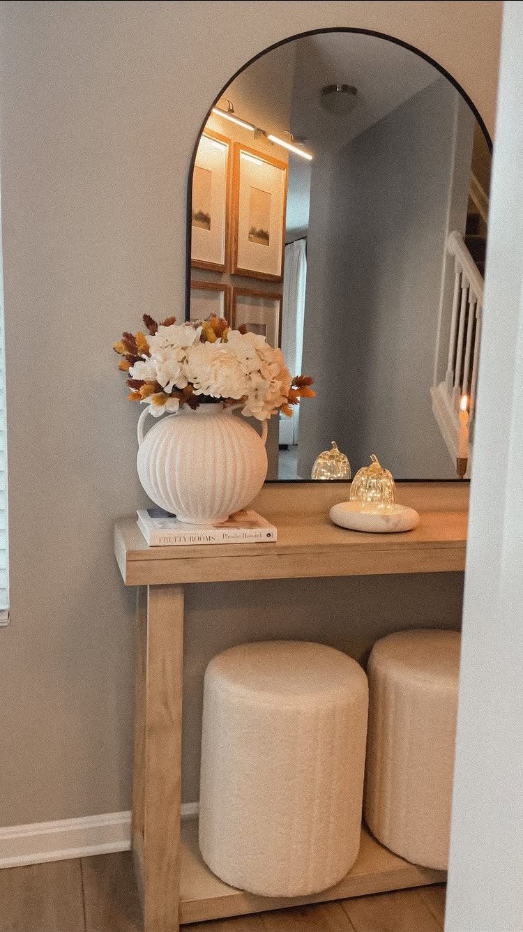 Slim light-wood entryway table with a white vase, arched mirror, and two ottomans.