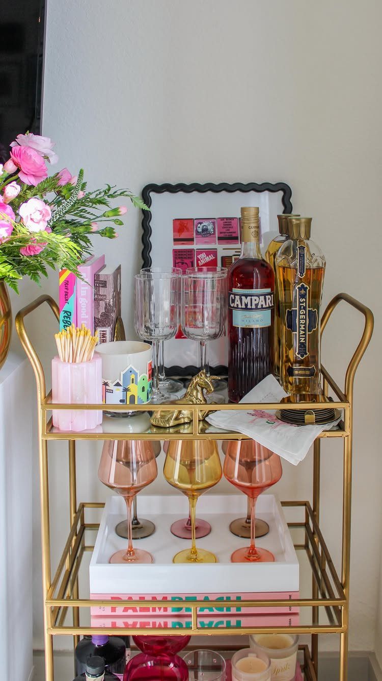 Gold metal bar cart stocked with liquor bottles, colorful glassware, and pink floral arrangements.