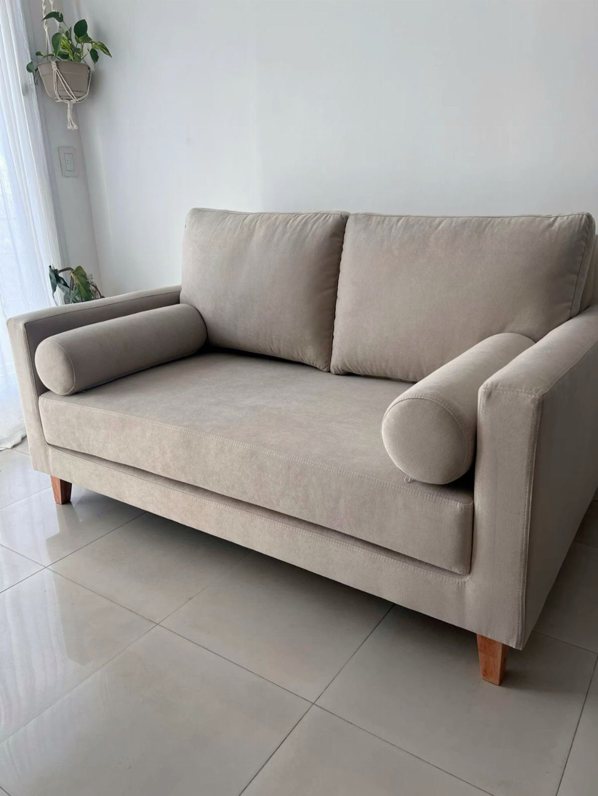 Beige fabric loveseat with bolster pillows on a light tiled floor.