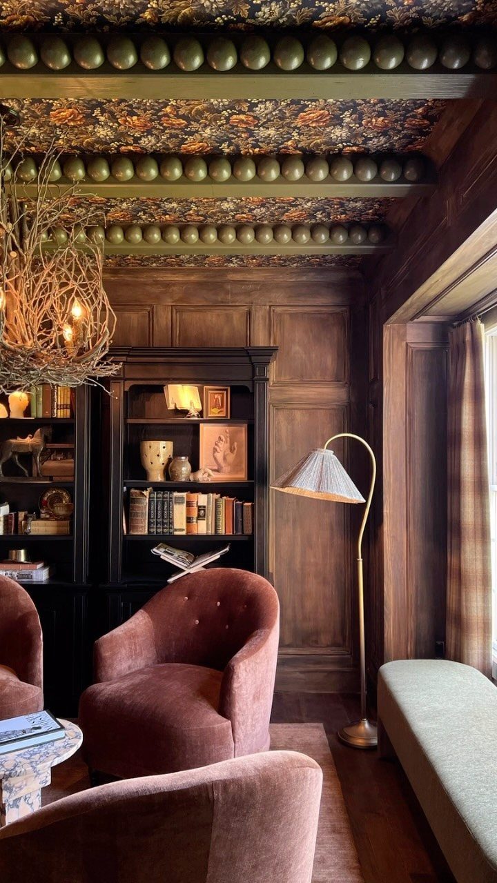 Floral wallpaper on a beamed ceiling in a wood-paneled living room with mauve chairs.