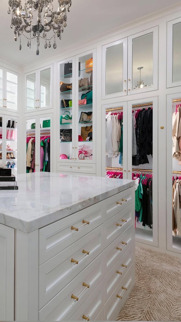 White walk-in closet with marble island and glass cabinet doors displaying clothing.