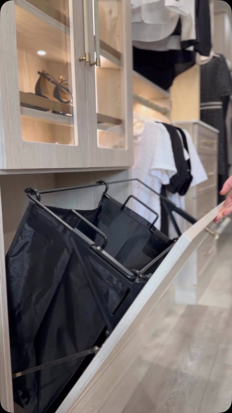 Tilt-out laundry hamper in a white closet with glass cabinets and hanging clothes.