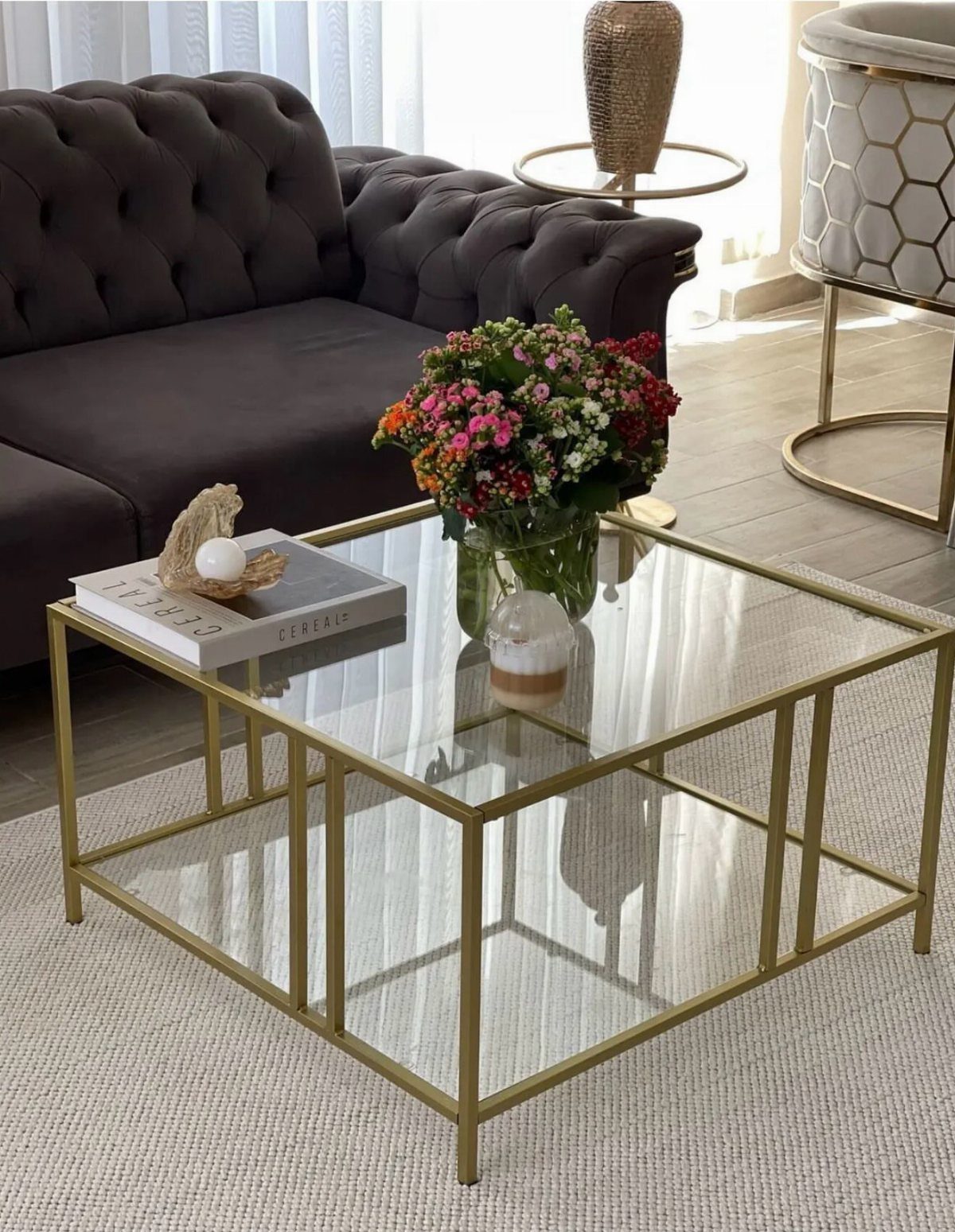Two-tiered gold and glass coffee table with flowers and a book on top.