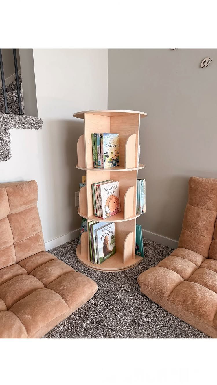 Circular wooden bookshelf with children's books placed in a room corner between tan chairs.