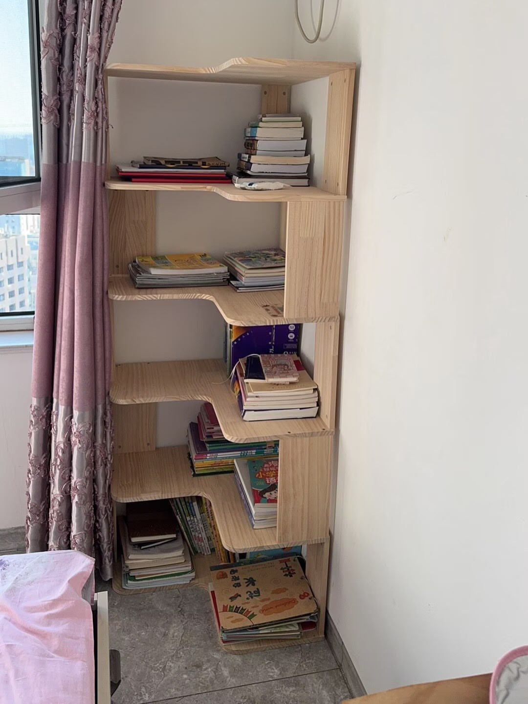Light wood corner bookshelf with curved shelves filled with books next to purple curtains.