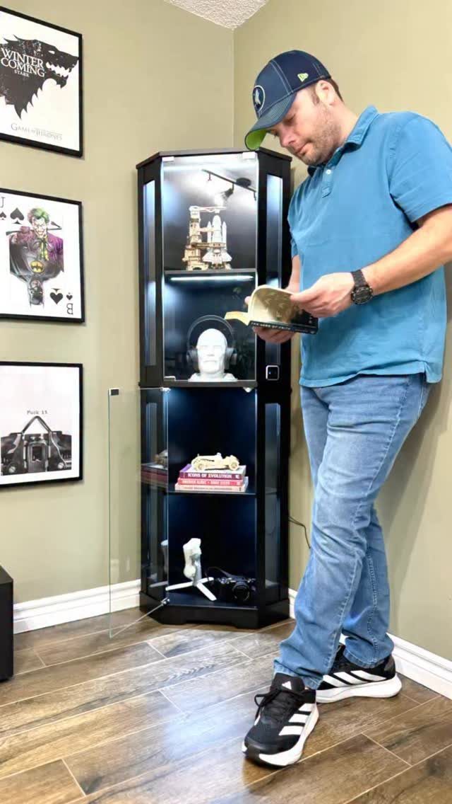 Man reading a book next to a black hexagonal glass corner bookshelf.