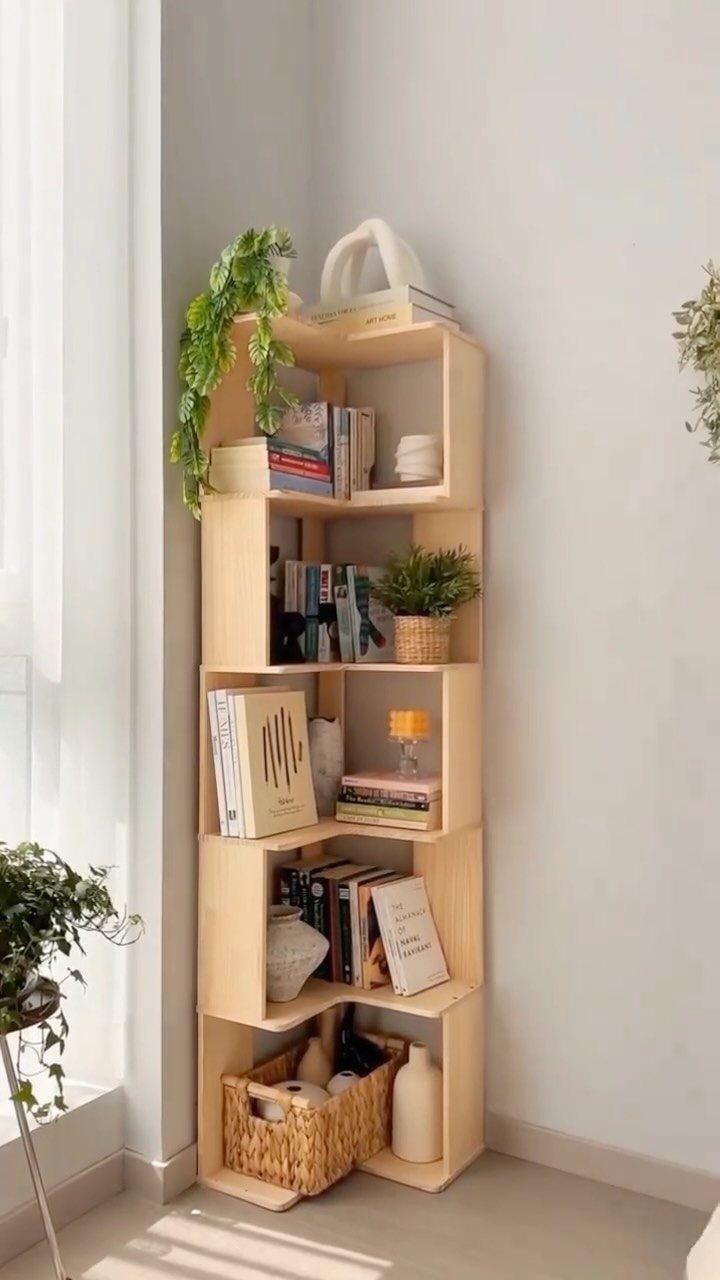 Tall light wood corner bookshelf filled with books and plants in a bright room.