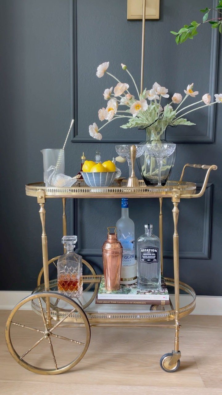 Gold bar cart with liquor bottles and white flowers against a grey wall.