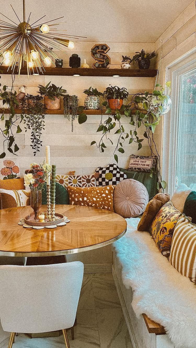 Dining nook with a wooden table, plush pillows, and shelves of potted plants.