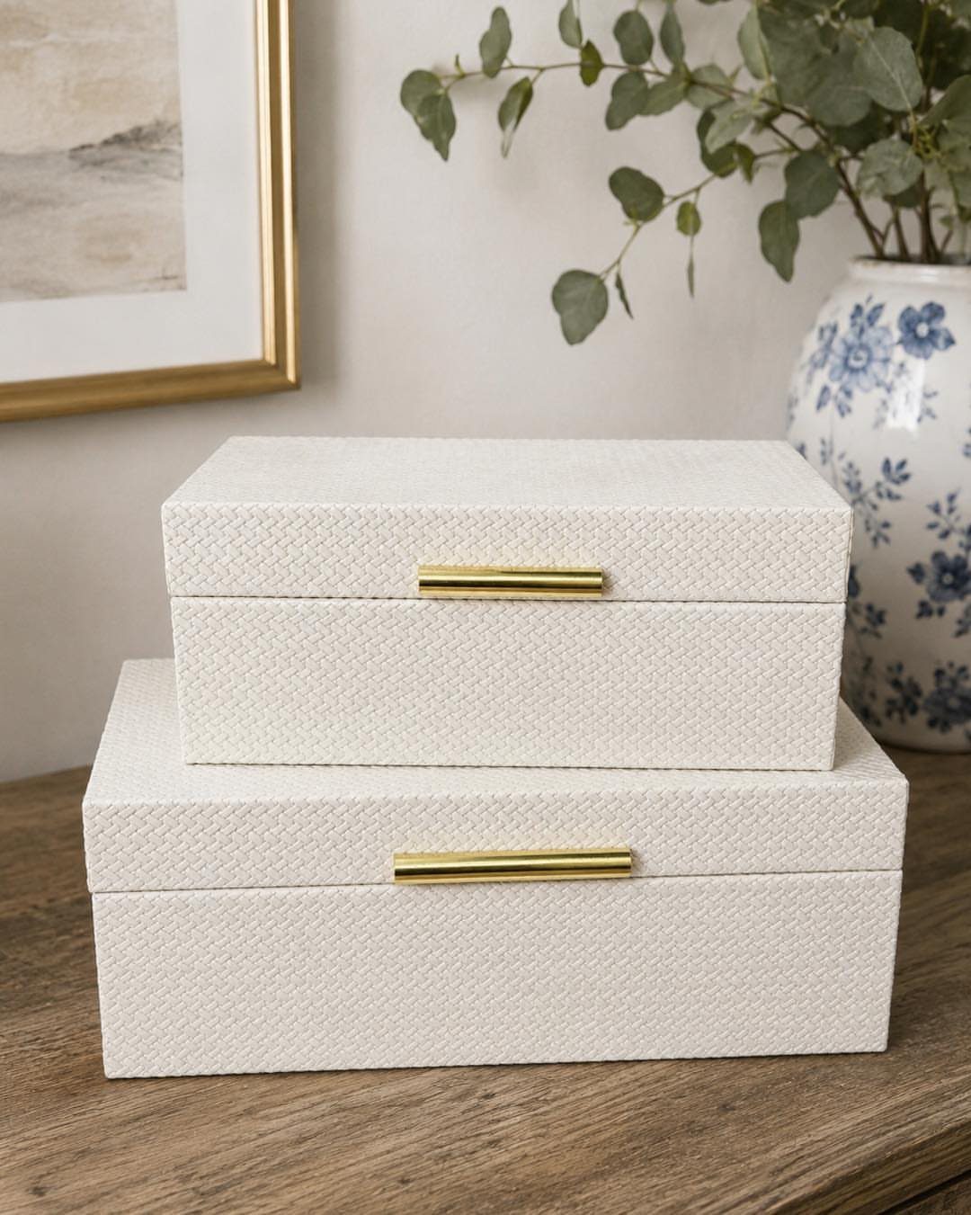 Stacked white textured decorative boxes with gold handles on a wooden dresser surface.