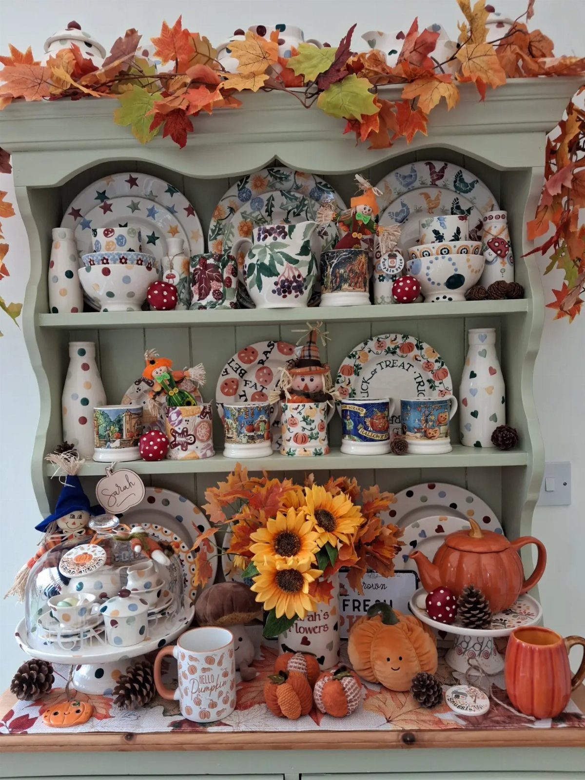 Autumnal decor with sunflowers and pumpkins arranged around ceramic dishes in a green hutch.