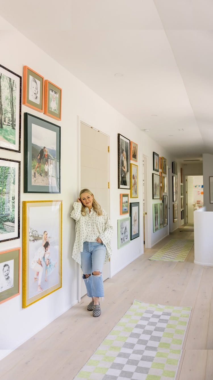 Woman leaning against a white hallway wall featuring a colorful gallery of framed photos.