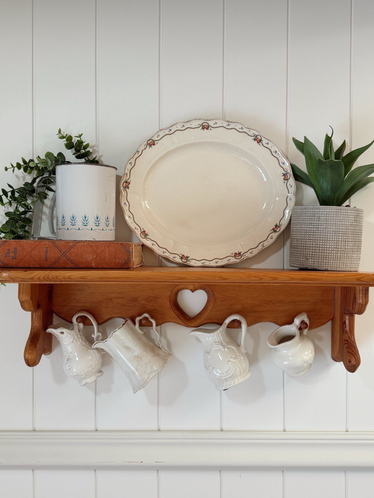 Wooden wall shelf with decorative pitchers, a platter, and a plant on white paneling.
