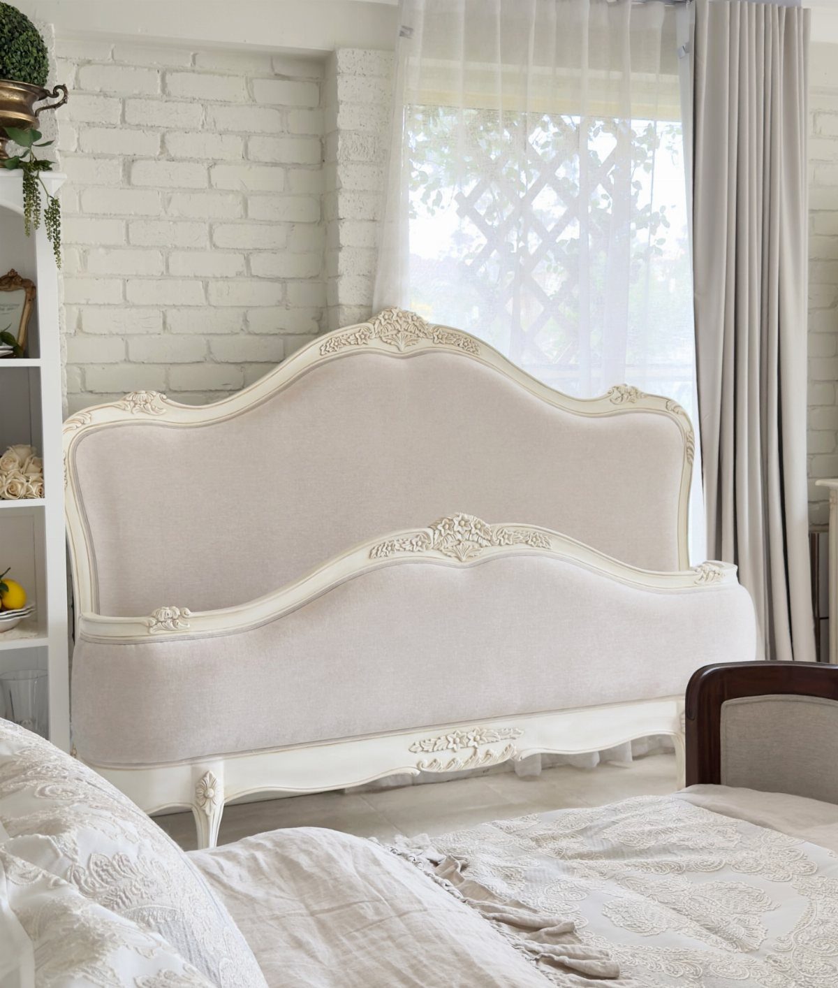 Ornate beige upholstered bed with carved white wooden frame in a bright room