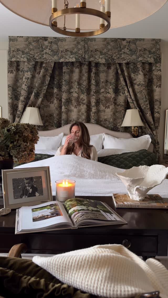 Woman in a bed with layered linens and floral curtains in a bedroom.
