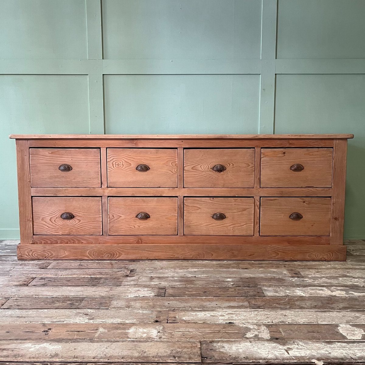 Rustic wooden dresser with eight drawers against a light green paneled wall.