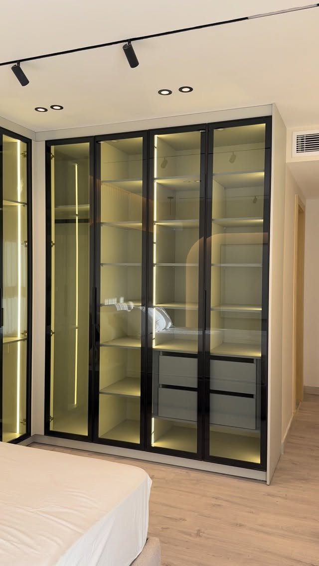 Black-framed glass wardrobe with interior lighting in a modern bedroom with light wood floors