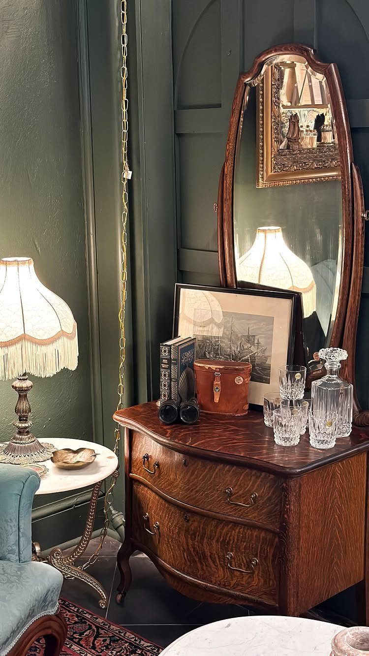 Antique wooden dresser with mirror, crystal glassware, and a fringed lamp in a room.