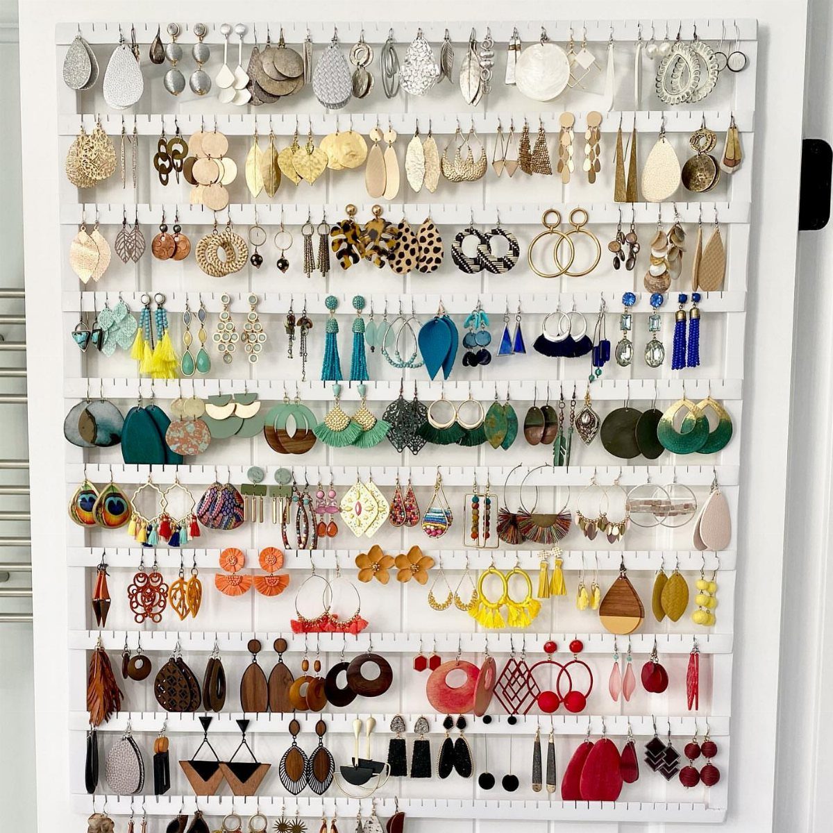 Colorful earrings hanging on a white wall-mounted organizer with multiple horizontal bars.