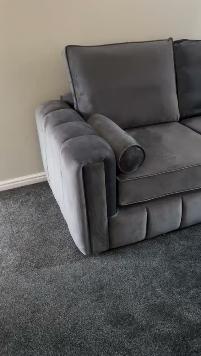 Grey velvet sofa with a bolster pillow on a dark grey carpet.