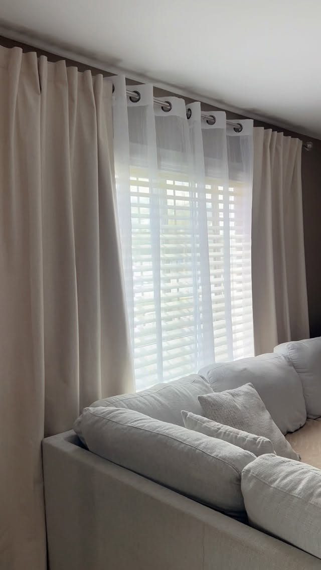 Beige and white sheer curtains hanging over a window above a grey sofa.