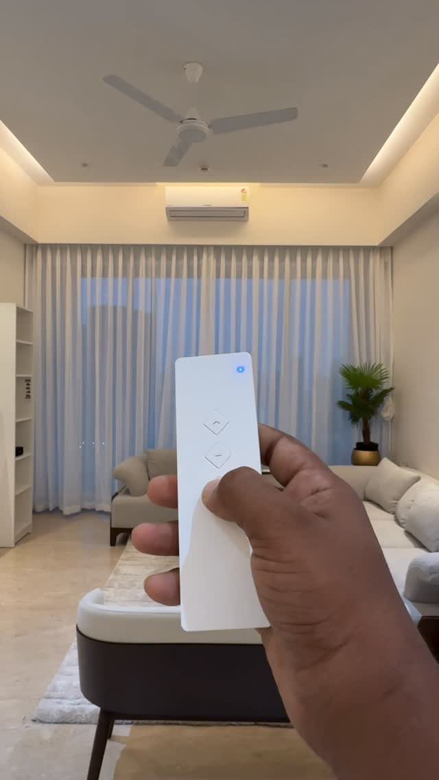 White remote control held in front of floor-to-ceiling white curtains in a living room.