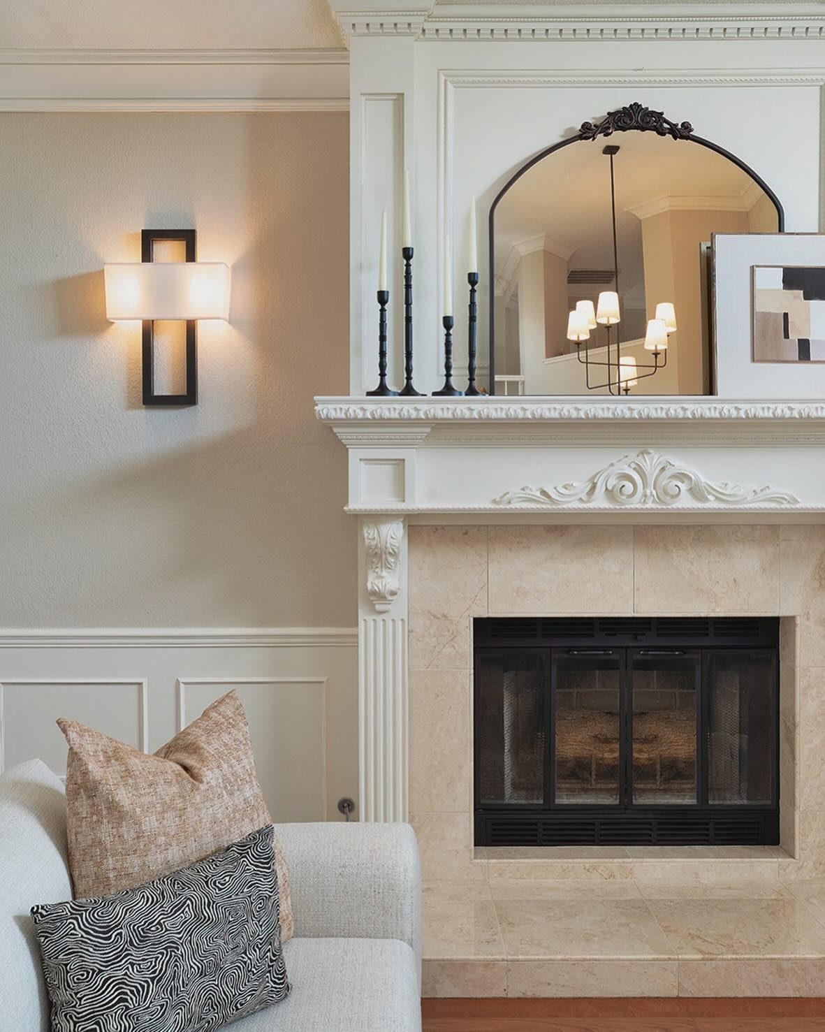 Arched mirror resting on a white fireplace mantel with black candlesticks and stone surround.