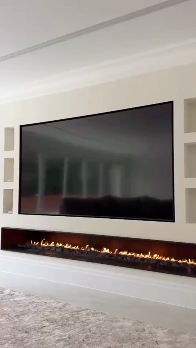 Linear gas fireplace with orange flames below a large wall-mounted television in white room.
