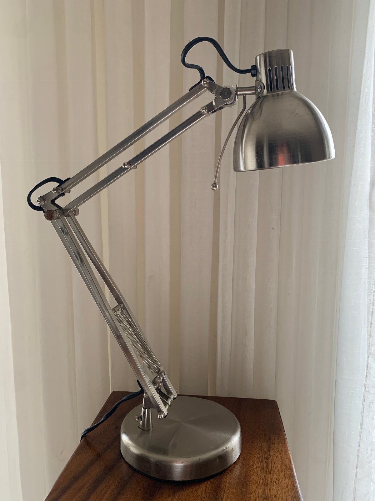 Silver metal adjustable arm desk lamp on a wooden table against white curtains.