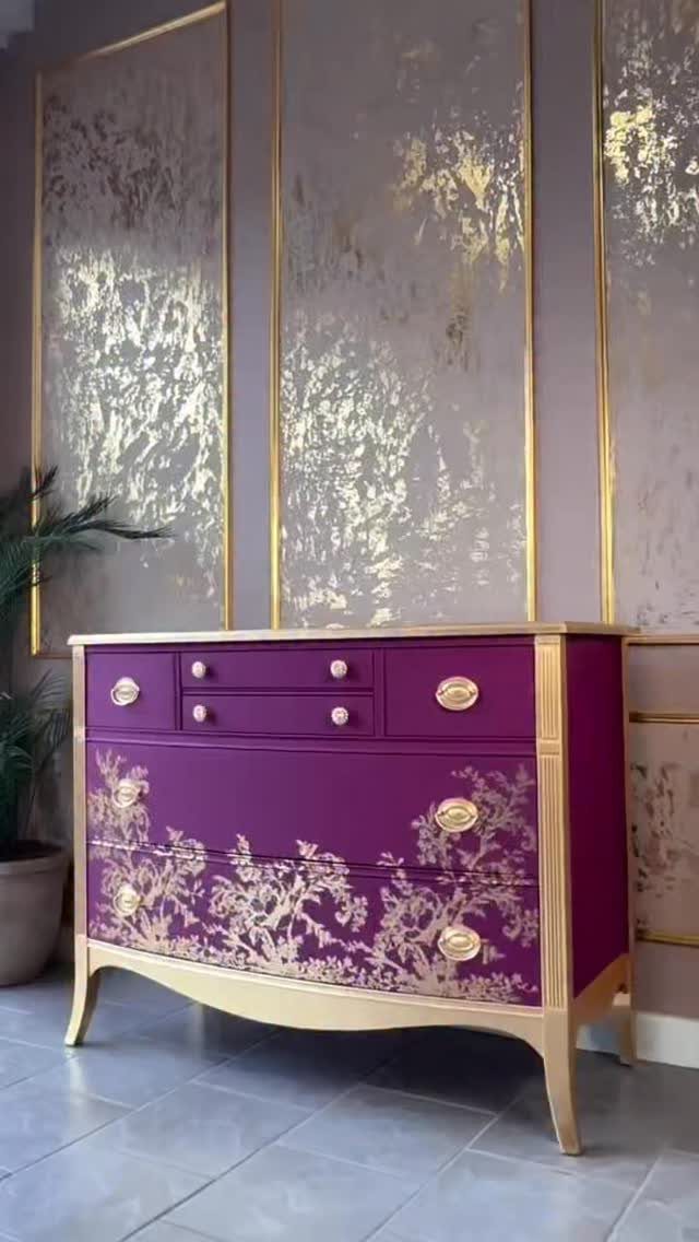 Purple dresser with gold ornate patterns and gold hardware against a metallic wall.