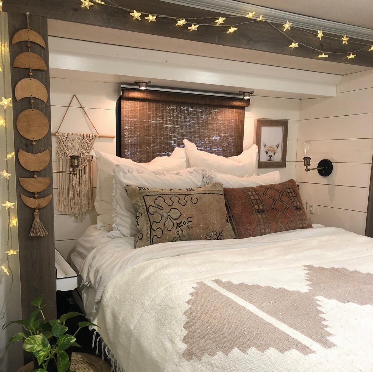 Cozy RV bedroom with white shiplap walls, patterned pillows, and hanging string lights.