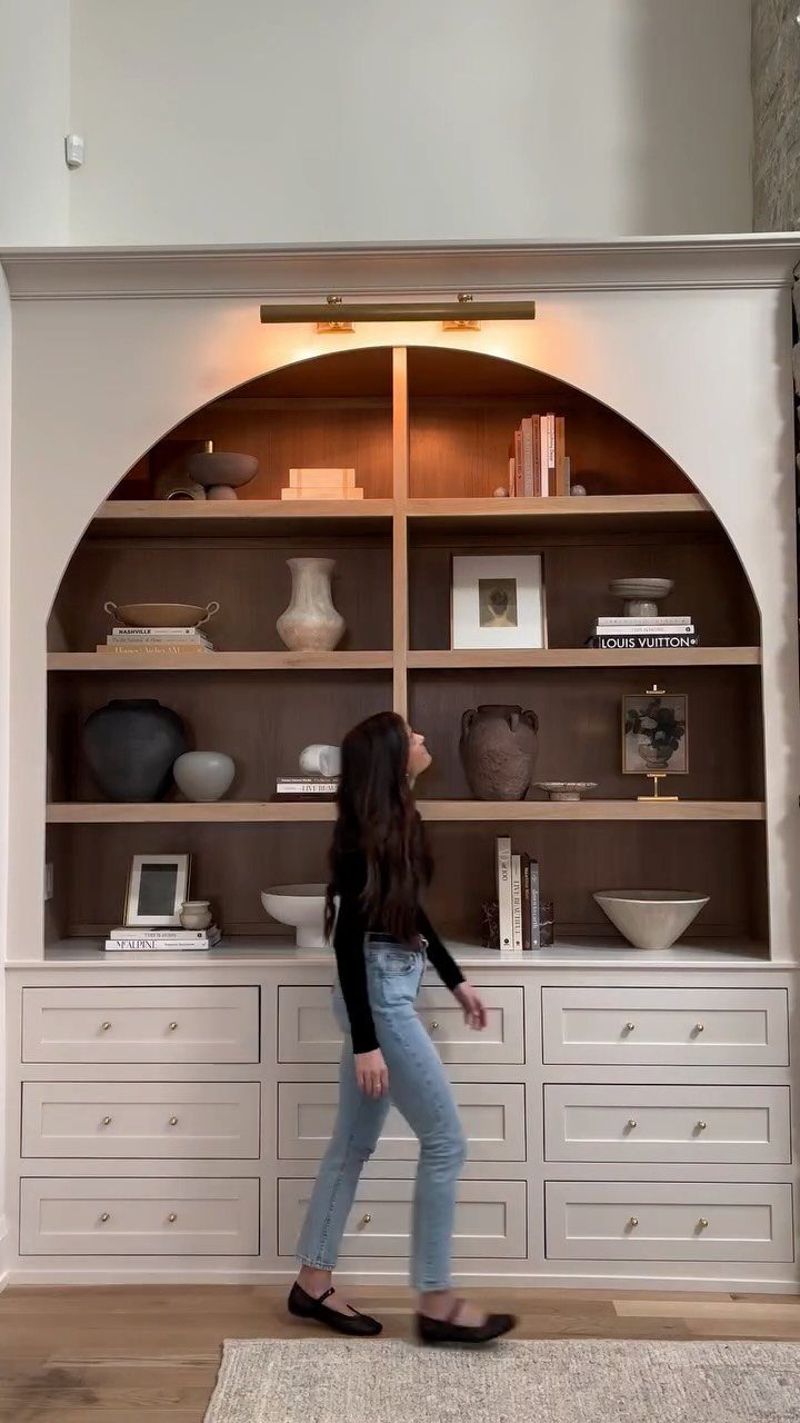 Arched white built-in shelves with spaced-out neutral decorative objects and a woman standing nearby.