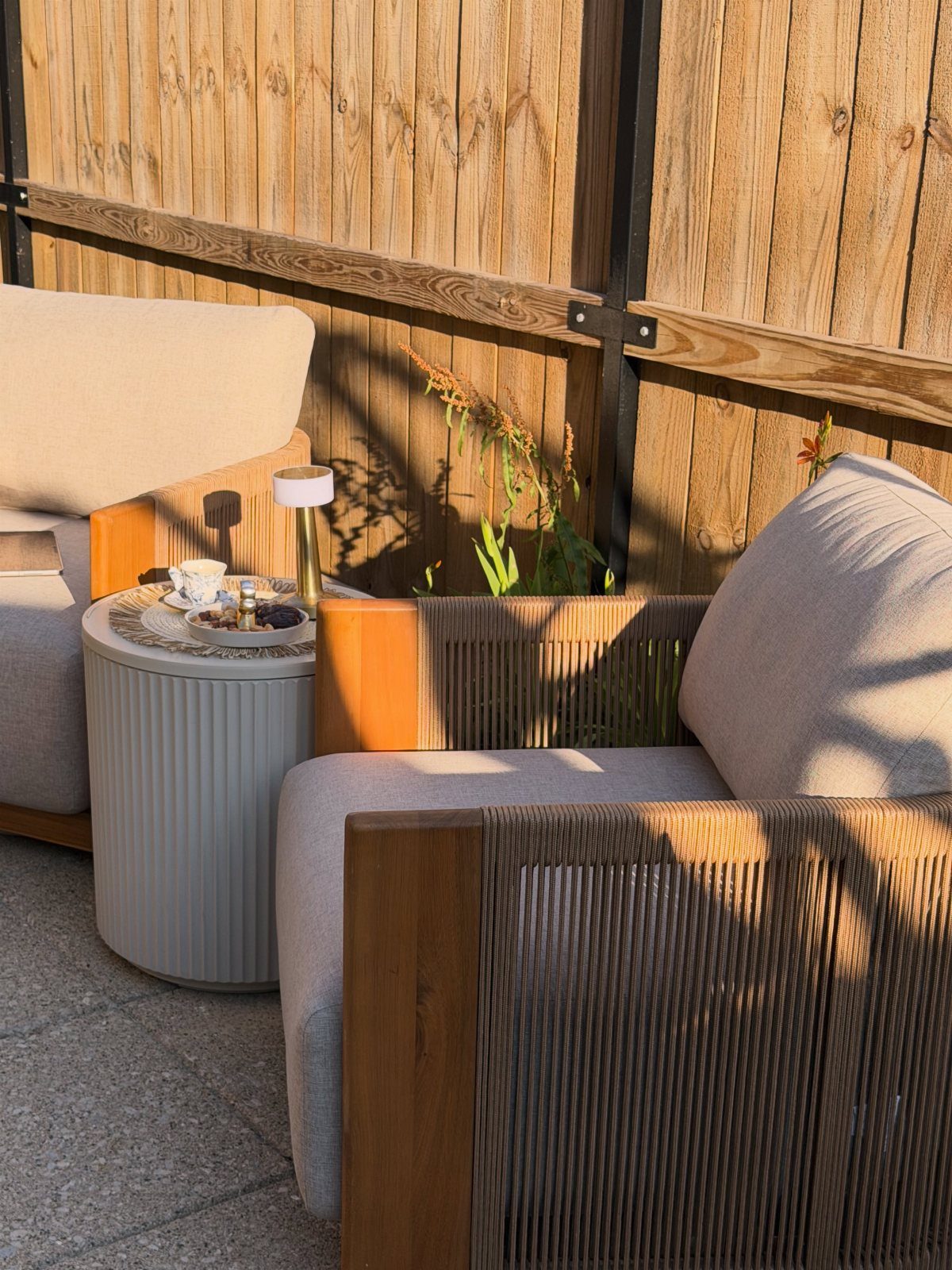 Small round grey outdoor coffee table between two modern lounge chairs by a fence