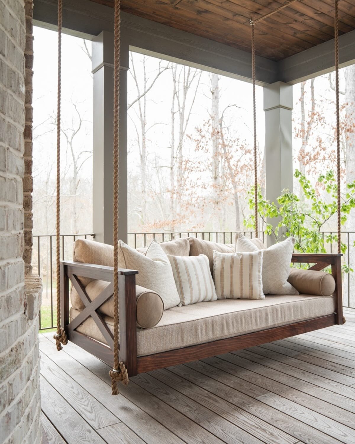 Wooden porch swing loveseat with beige cushions and pillows on a grey deck.