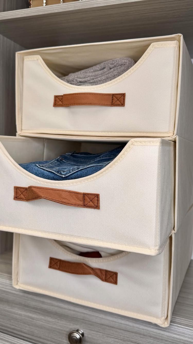 Cream fabric storage bins with leather handles holding folded clothes on a shelf.