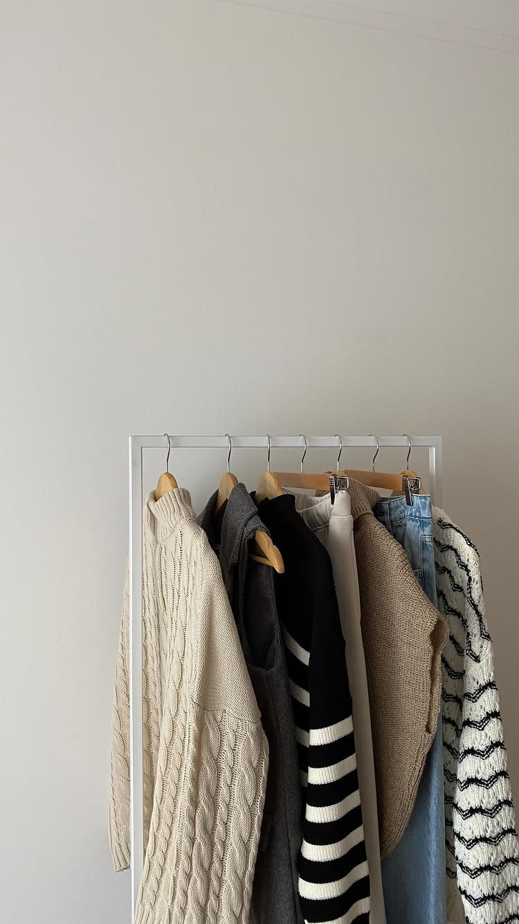 Cream and grey sweaters hanging on a white metal clothing rack against a wall.