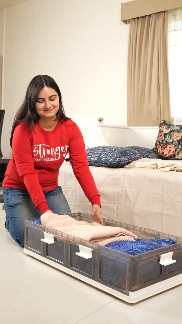 Woman placing a folded beige sweater into a large clear plastic storage bin.