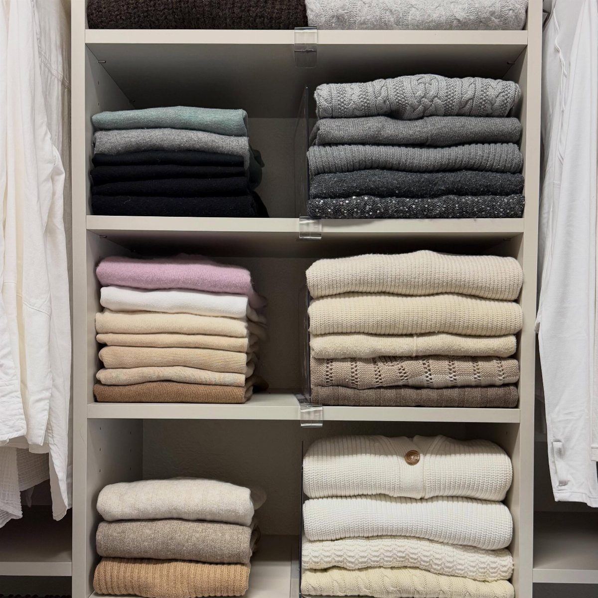 Folded sweaters in neutral tones organized on white shelves with clear shelf stackers.