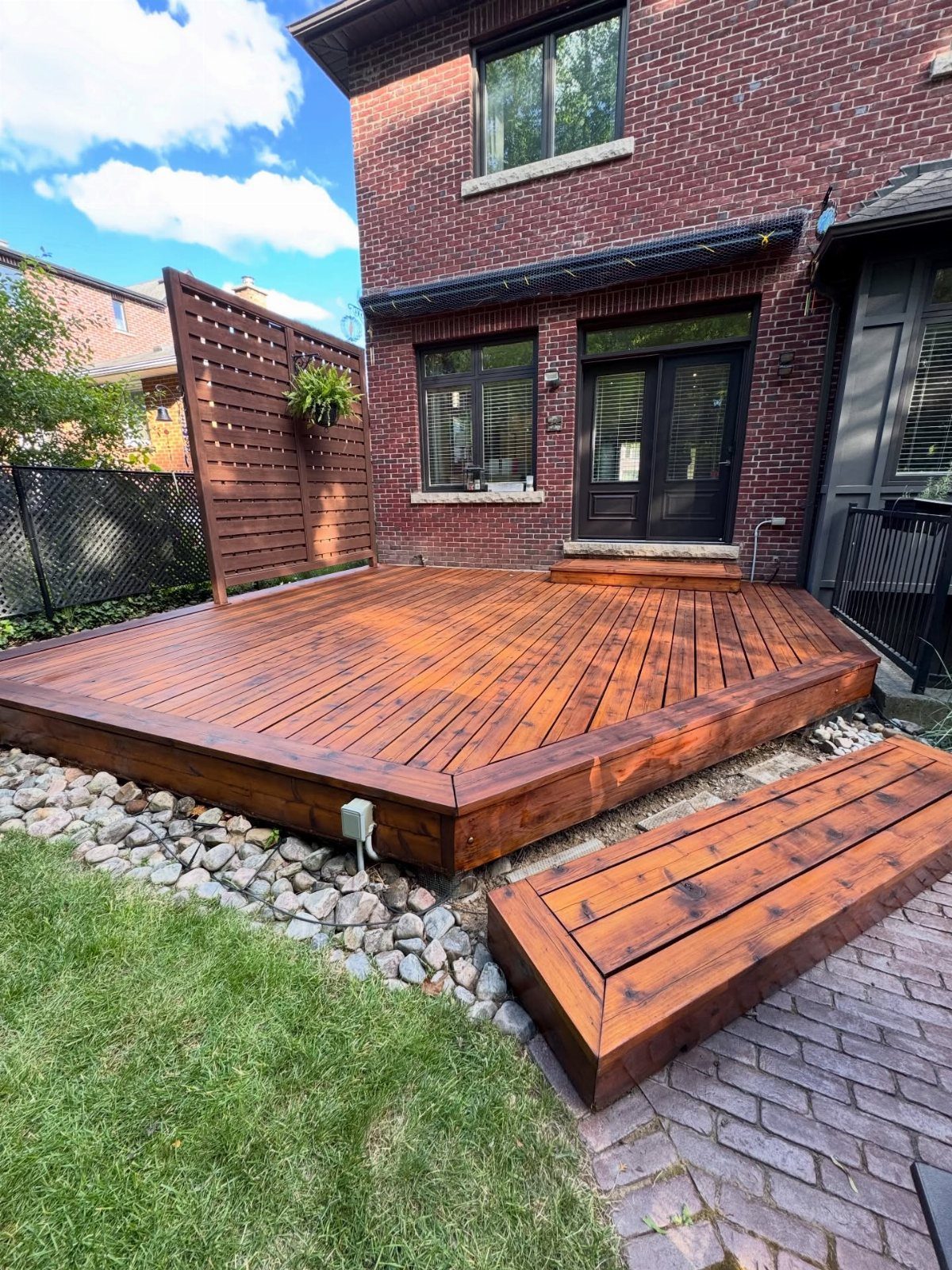 Reddish-brown stained wooden deck and bench outside a brick house with green grass.