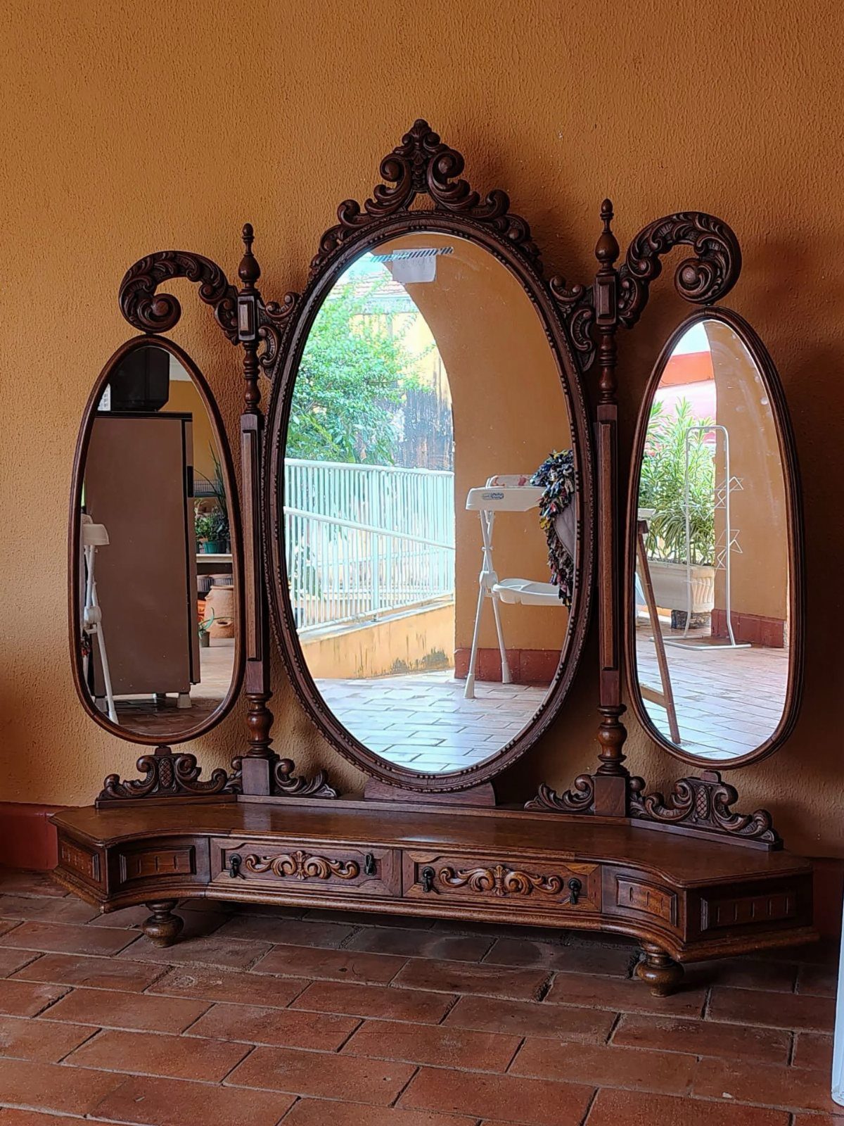 Ornate dark wood vintage vanity with three oval mirrors against a yellow wall.